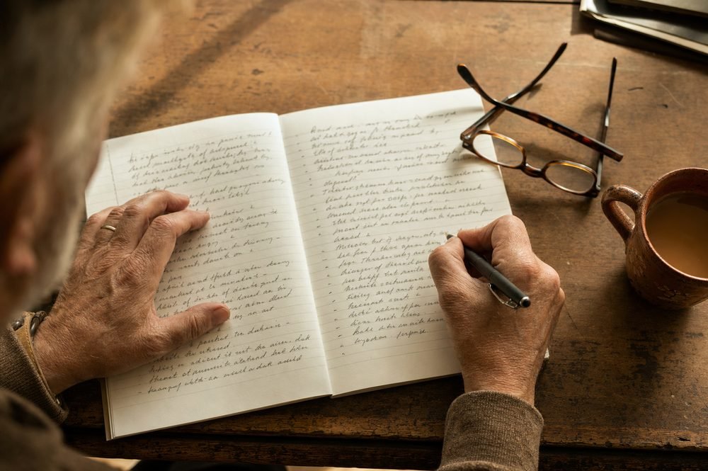Hands writing a long list in a notebook on a wooden desk, reading glasses and a mug of tea beside it